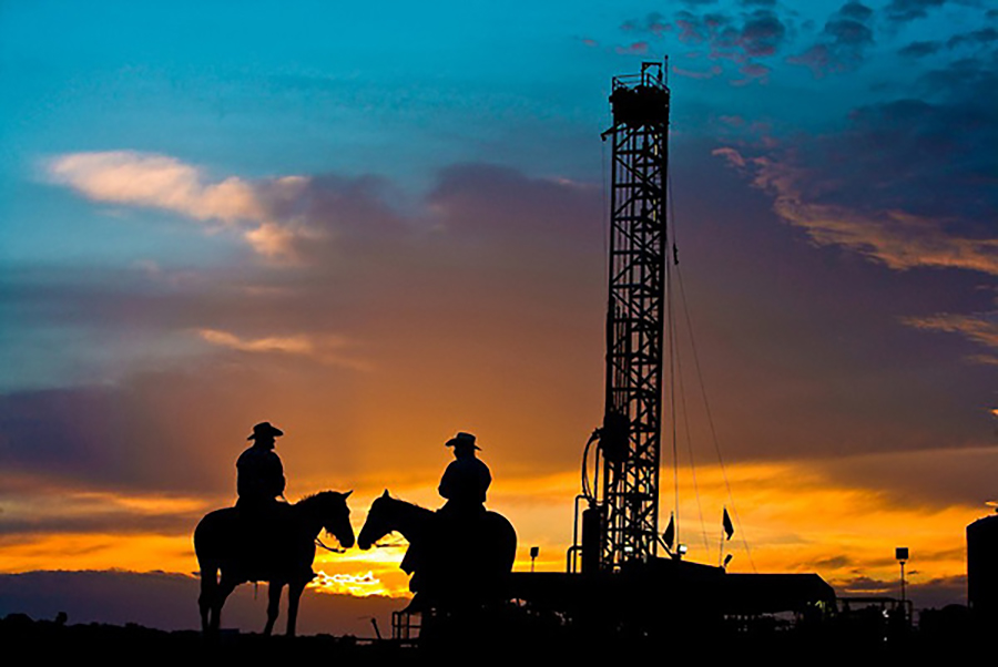 Two cowboys on horseback in front of an oil and gas drilling rig in the Eagle Ford Shale play at sunset in South Texas